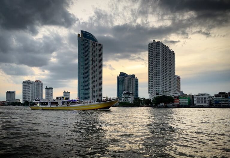 Boat cruising along the Chao Phraya River with a backdrop of modern skyscrapers in Bangkok at sunset.