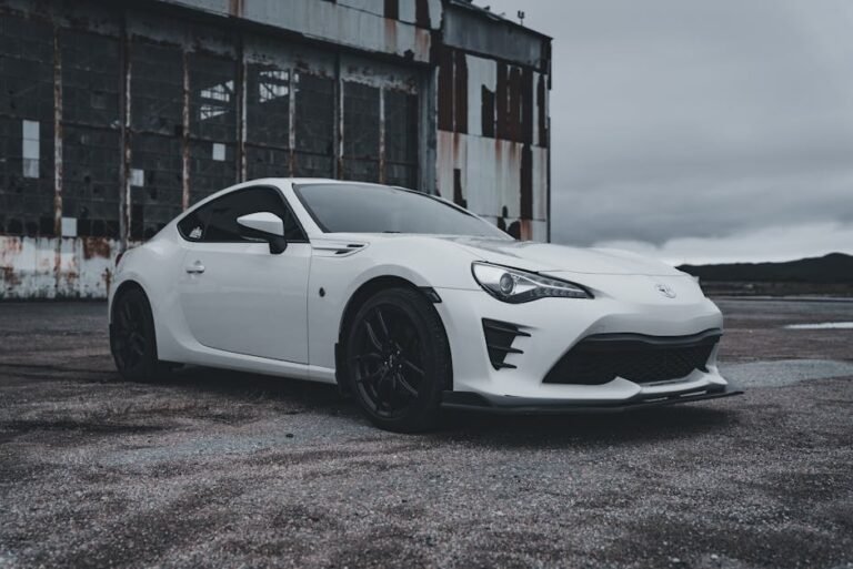 White sports car parked by an abandoned warehouse on an overcast day.
