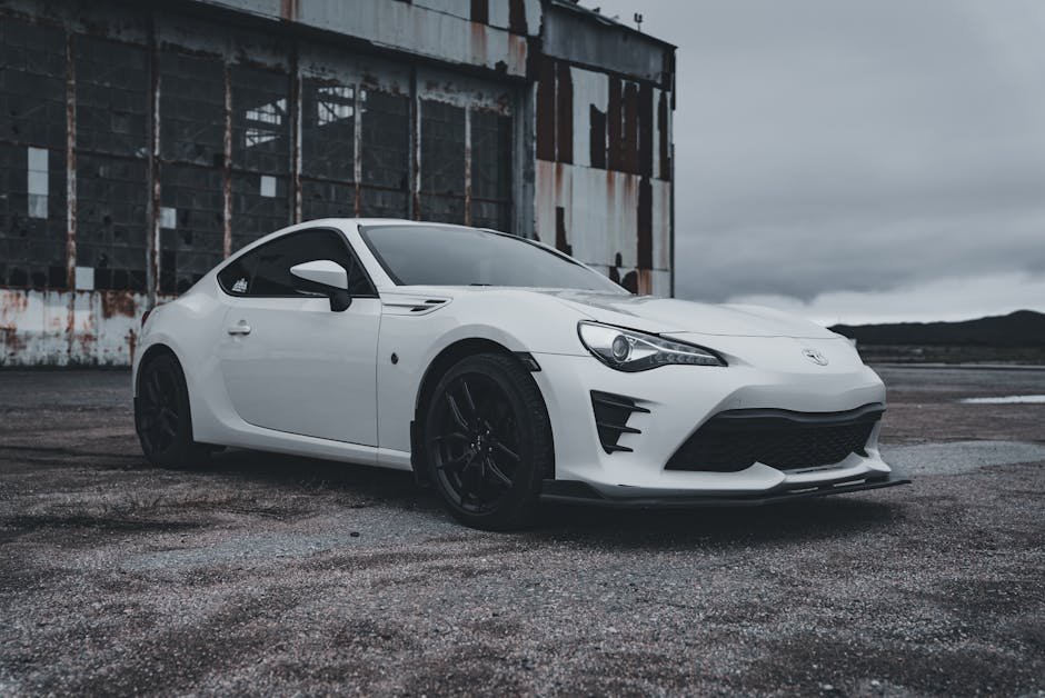 White sports car parked by an abandoned warehouse on an overcast day.