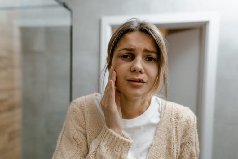 A woman indoors with a worried expression, touching her cheek, in a light-toned room.