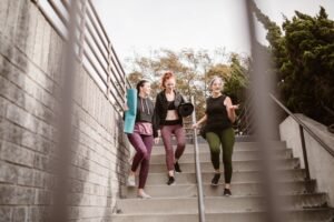 Three women walking down steps outdoors, carrying yoga mats, enjoying a fitness conversation.