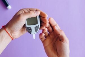 Hands holding a glucometer to test blood sugar levels on a purple background.