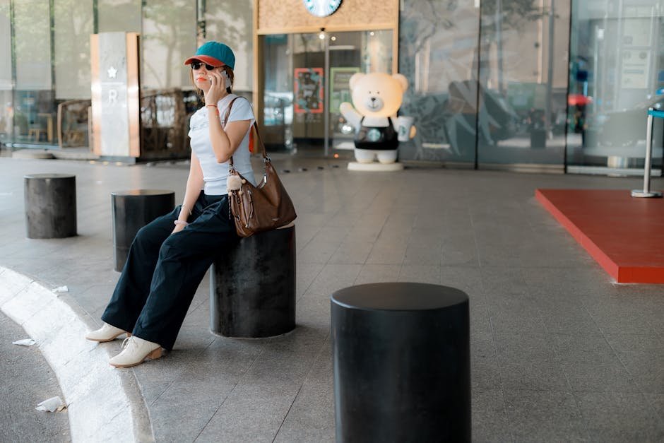 Stylish woman sitting on street furniture, talking on phone outside modern building.