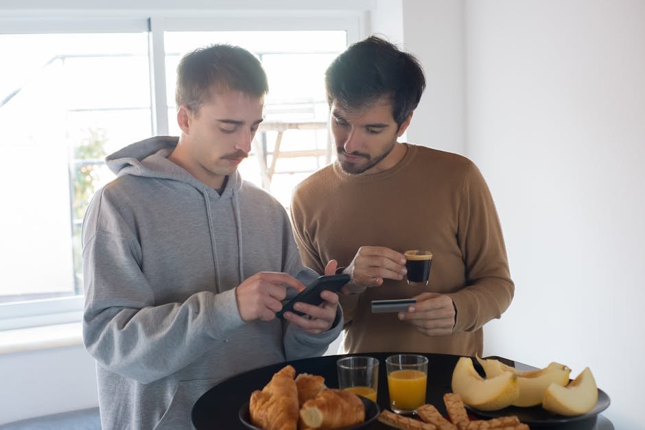 Two men having breakfast with coffee and fruit indoors, engaging with smartphone and credit card