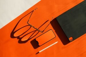 Glasses, smartphone, tablet, and stylus arranged on an orange background with shadows.