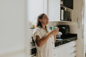 Relaxed woman standing in a kitchen, smiling and holding a cup of coffee, enjoying a sunny morning.