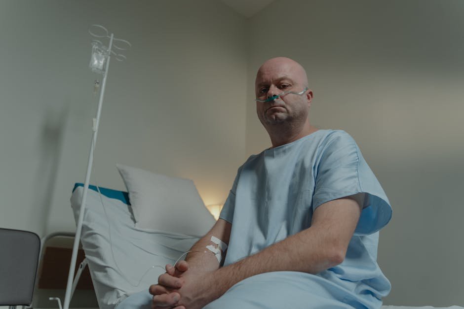 Bald man in hospital gown sitting on bed with nasal cannula and IV in a clinical setting.